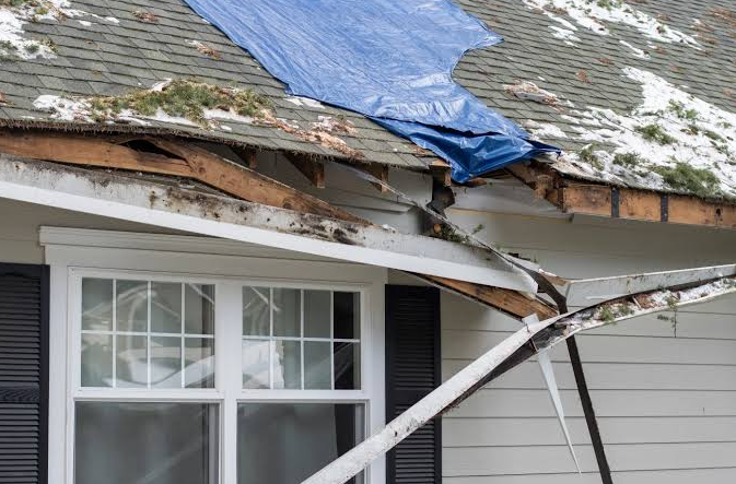 Storm-damaged roof with missing shingles, exposed wood framing and a blue tarp covering the damage